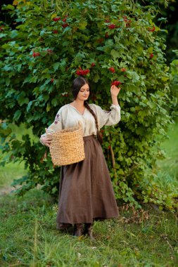 Geleneksel Ukrayna kıyafetleriyle uzun siyah örgülü güzel esmer kadın yeşil arka planda parlak kırmızı viburnum ile poz veriyor, çarpıcı bir portre, canlı bir açık hava sahnesi..