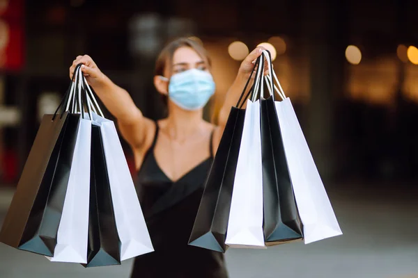 センターモールの近くにショッピングバッグを保持保護医療マスクの黒のドレスのスタイリッシュな女性 コロナウイルスの流行で買い物 消費主義 ブラックフライデーの概念 — ストック写真