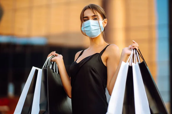 モールの近くにショッピングバッグを保持保護医療マスクのスタイリッシュな女性 コロナウイルスの流行で買い物 消費主義 ブラックフライデーの概念 — ストック写真