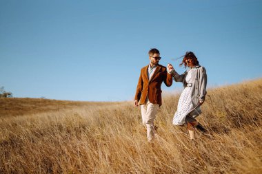 Güz tarlasında sonbahar havasının tadını çıkaran şık genç bir çift. Tarlada yürüyen ve kucaklaşan sevimli bir çift. Gençlik, aşk ve yaşam tarzı kavramı.