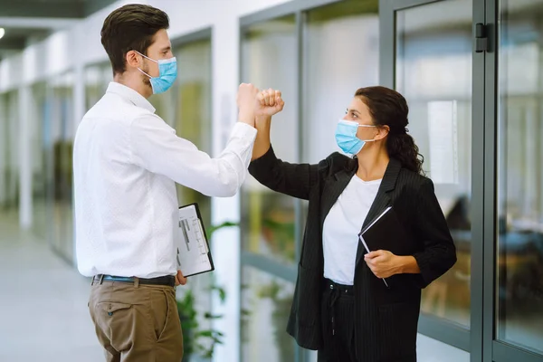 Business colleagues in protective face masks in modern office  bumping elbows while greeting each other at work. Businesspeople back at work in office after quarantine. Covid-19.