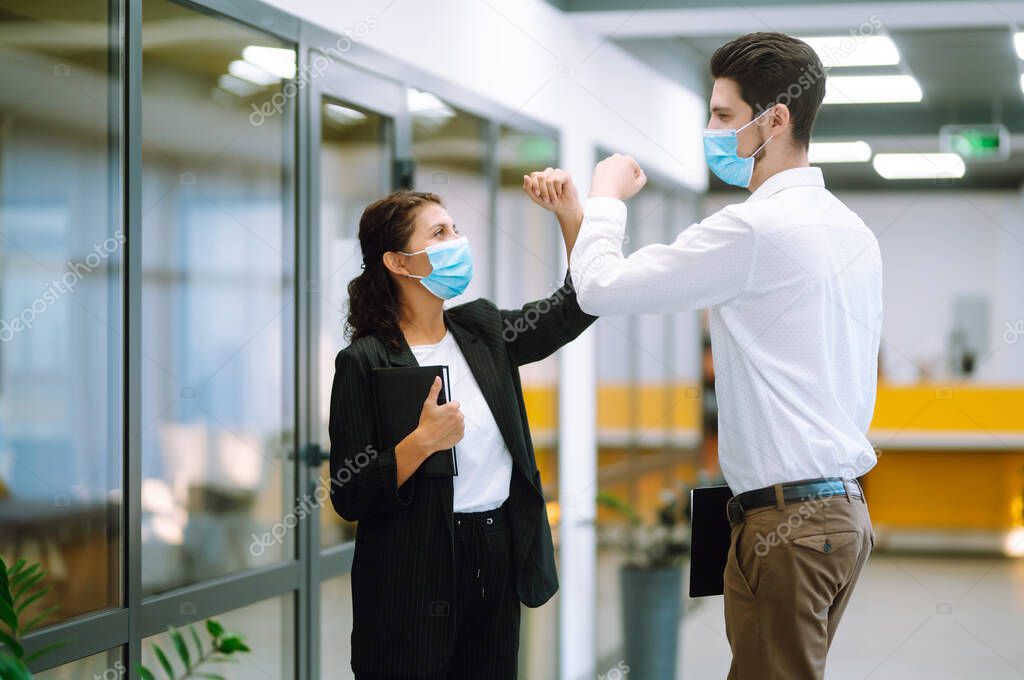 Business colleagues in protective face masks in modern office  bumping elbows while greeting each other at work. Businesspeople back at work in office after quarantine. Covid-19. 