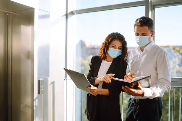 Two colleagues in protective face masks in modern office discussing together work issues standing near elevator. Business People back at work in office after quarantine. Covid-19.