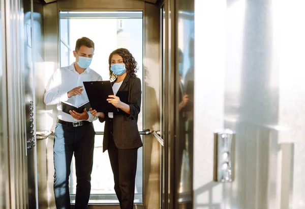 Business colleagues in medical face mask talking while standing in elevator at modern office. Boss and secretary or assistant work as team.  Business partners back at work in office after quarantine.