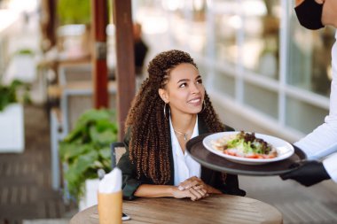 Korunaklı maske ve eldiven takan genç garson Coronavirus salgını sırasında bir müşteriye yemek getirirken. Garson lokantada görev başında..