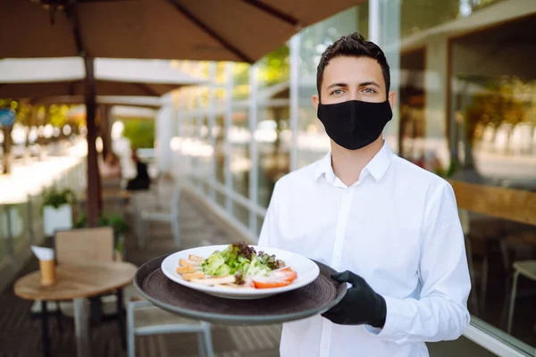 彼の顔に保護マスクの若いウエイターとカフェで食べ物とプレートを保持手袋 レストランでの勤務中にウェイターが働いています Covid 2019 — ストック写真