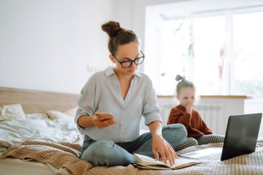 Genç anne cep telefonuyla konuşuyor ve dizüstü bilgisayarla çalışıyor. İş kadını evde küçük bir kızla çalışıyor. Doğum izninde çalış. Covid 19.
