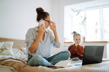 Genç anne cep telefonuyla konuşuyor ve dizüstü bilgisayarla çalışıyor. İş kadını evde küçük bir kızla çalışıyor. Doğum izninde çalış. Covid 19.