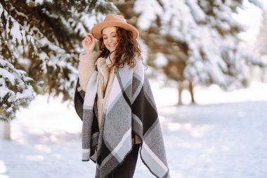 Güzel bir kadın karlı ağaçların arasında duruyor ve ilk karın tadını çıkarıyor. Şapka, ekose atkı ve palto giyen genç bir kadın. Kış tatilin kutlu olsun. Noel.