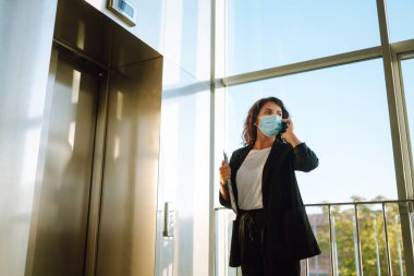 Modern ofisteki asansörün yanındaki virüs salgını sırasında cep telefonuyla iletişim kuran iş kadını. Covid 19.