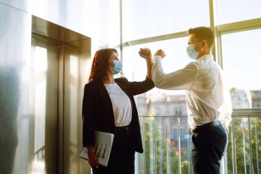 Koruyucu yüz maskesi takmış iş arkadaşları modern ofisteki asansörün yanında birbirini selamlarken dirseklerini çarpıyorlar. Covid 19.