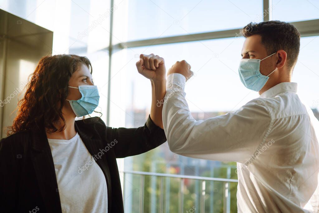 Business colleagues in protective face masks bumping elbows while greeting each other near elevator at modern office. Covid-19.