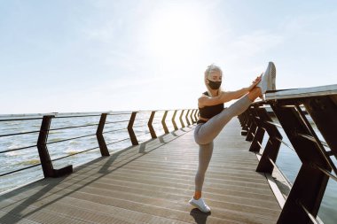Sahil kenarında koruyucu maske takan sağlıklı bir kadın. Spor kıyafetli genç bir kadın gün doğumunda plajda egzersiz yapmadan önce bacak esnetiyor. Sabah sporu. Covid 19.