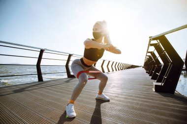 Sahil kenarında koruyucu maske takan sağlıklı bir kadın. Spor kıyafetli genç bir kadın gün doğumunda plajda egzersiz yapmadan önce bacak esnetiyor. Sabah sporu. Covid 19.