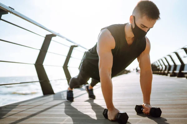 Handsome muscular man in protective mask, sports outfit  doing functional exercises on seaside promenade.  Sportsman performing bicep curl with dumbbells. Morning workout. Covid-19.