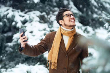Ceketli, kazaklı, atkılı ve gözlüklü şık bir adam karlı bir parkta selfie çekiyor. İş adamı bir telefon kullanır ve karlı bir ormanda kış anlarının tadını çıkarır. Kar yağışı. Video çağrısı.