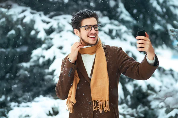 Stylish man in a coat,  sweater, scarf and glasses takes a selfie  in a snowy park. Businessman uses a phone and  enjoying winter moments in a snowy forest. Snowfall. Video call.