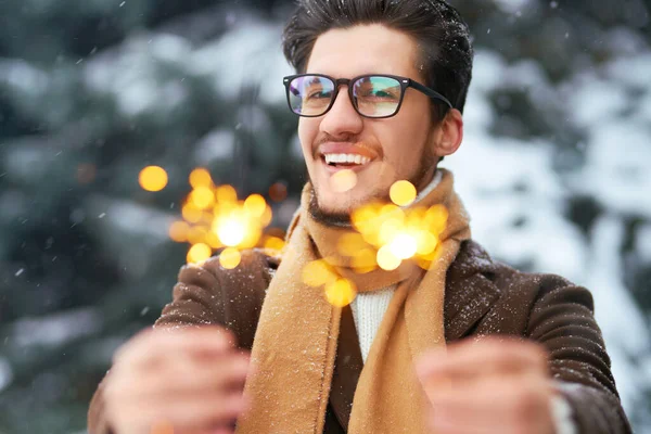 Sparklers in the hands of a young man. Stylish man in a coat,  sweater, scarf and glasses celebrates winter holidays. Christmas, New year. Winter lifestyle.