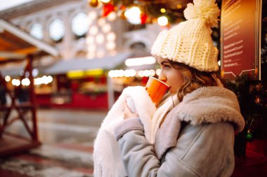 Noel pazarında yürürken sıcak kahve içen genç bir kadın. Akşamları Noel ışıklarıyla süslenmiş. Noel ve yeni yıl büyüsü. Işıklar yansın.