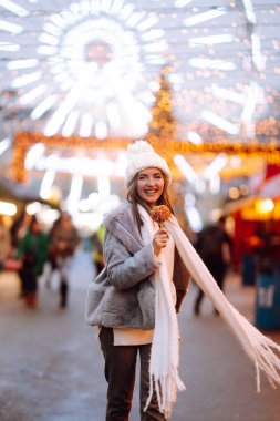 Noel pazarında karamelli elmalı mutlu bir kadın. Noel ve Yeni Yıl 'da açık hava aktiviteleri. Genç bir kadın kış anlarından zevk alıyor. Işıklar yansın.
