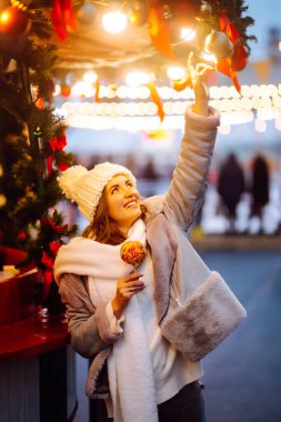 Noel pazarında karamelli elmalı mutlu bir kadın. Noel ve Yeni Yıl 'da açık hava aktiviteleri. Genç bir kadın kış anlarından zevk alıyor. Işıklar yansın.