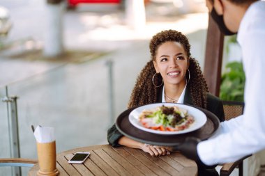 Koruyucu maskeli genç garson ve sipariş edilmiş yemeklerle birlikte eldivenler hazır. Coronavirus salgını sırasında kafede garsonmuş. Covid 2019.