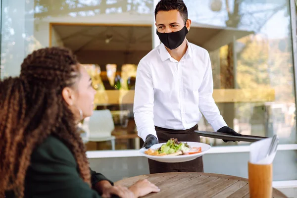 保護顔のマスクと手袋で若いウエイターは ゲストにサービスを提供する準備ができて 食事を注文した コロナウイルス発生中にカフェでウェイター Covid 2019 — ストック写真