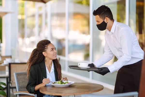 Young waiter in protective face mask and gloves with ordered meals, ready to serving guest. Waiter in cafe during coronavirus outbreak. Covid-2019.