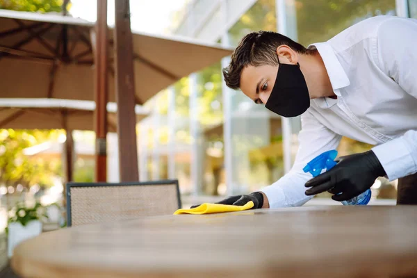 Young Waiter in protective face mask and gloves sanitizing surfaces, cleaning the table with disinfectant spray in a restaurant. Covid- 2019.