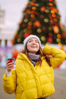Yeni yıl ağacının yanında yürürken sıcak kahve içen genç bir kadın. Mutlu kadın örgü şapka ve eşarp takıyor, soğuk kış gününde termosla sıcak çay tutuyor. Noel şenliği, kış tatili 