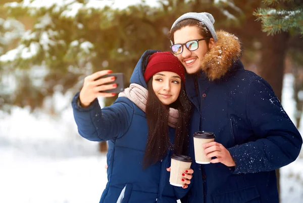 Happy couple with coffee in winter forest make selfie. The man and the woman have a rest in the winter park. Happy winter time. Christmas.