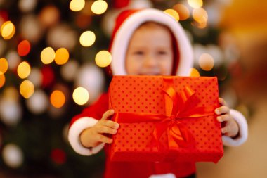 Küçük çocuğun elinde Noel hediyesi. Noel Baba kostümlü çocuk Noel ışıklarının ve ağacın arkasında kırmızı hediye kutusunu tutuyor. Kış tatili, yeni yıl.