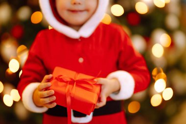 Küçük çocuğun elinde Noel hediyesi. Noel Baba kostümlü çocuk Noel ışıklarının ve ağacın arkasında kırmızı hediye kutusunu tutuyor. Kış tatili, yeni yıl.