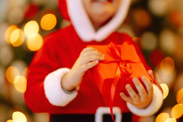 Christmas gift in the hand of Little child.  Kid in santa costume holding red present  box on the background of Christmas lights and tree. Winter holiday, New Year.