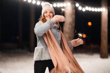 Noel 'in ışıltısı ellerde. Kış ormanında parıltı tutan neşeli genç bir kadın. Şenlikli çelenk ışıkları. Noel, yeni yıl.
