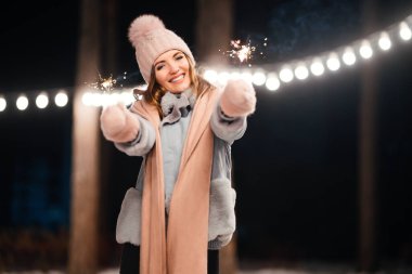 Noel 'in ışıltısı ellerde. Kış ormanında parıltı tutan neşeli genç bir kadın. Şenlikli çelenk ışıkları. Noel, yeni yıl.