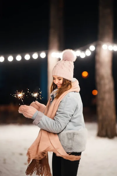 Christmas sparkles in hands. Cheerful young woman celebrating  holding sparkles in the winter forest. Festive garland lights. Christmas, new year.