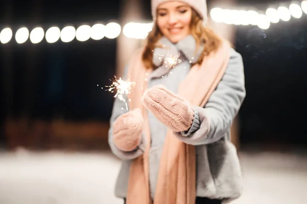 Christmas sparkles in hands. Cheerful young woman celebrating holding sparkles in the winter forest. Festive garland lights. Christmas, new year.