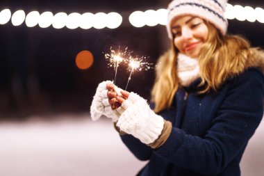 Yanan havai fişek kıvılcımını tutan kadın el. Genç bir kadın kış ormanında parıltı tutarak kutlama yapıyor. Şenlikli çelenk ışıkları. Noel, yeni yıl partisi.