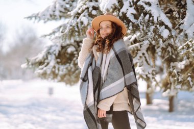 Şapkalı güzel bir kadın, ekose atkısı ve paltosu var. Kar ormanında neşe içinde poz veriyor. Karlı bir parkta kışın eğlenen neşeli kıvırcık kadın. Tatiller, sezon ve eğlence konsepti. Noel.