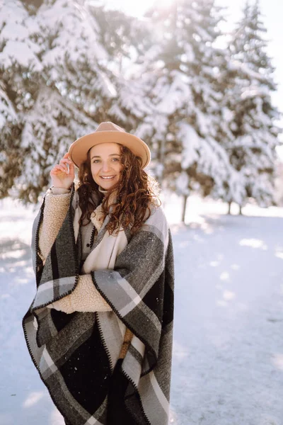 Beautiful woman in hat, plaid scarf and coat posing with joy outside in the snow forest. Cheerful curly lady enjoying winter moments in a snowy park. Holidays, season and leisure concept. Christmas.