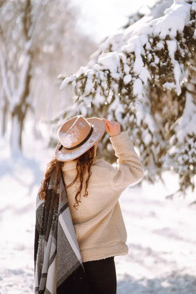 Beautiful woman in hat, plaid scarf and coat posing with joy outside in the snow forest. Cheerful curly lady enjoying winter moments in a snowy park. Holidays, season and leisure concept. Christmas.