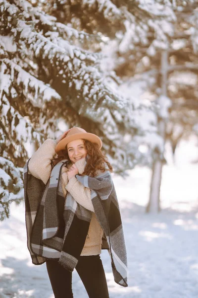 Beautiful woman in hat, plaid scarf and coat posing with joy outside in the snow forest. Cheerful curly lady enjoying winter moments in a snowy park. Holidays, season and leisure concept. Christmas.