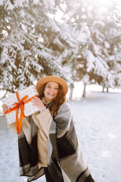 Big gift box with red ribbon in the woman hands.  Fashion young woman holding a Christmas present standing among snowy trees and enjoying first snow. Holidays, season and leisure concept.