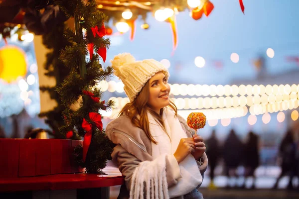 クリスマスマーケットでキャラメルアップルと若い女性 お祝いの通りの市場でポーズ冬のスタイルの服の笑顔の女性 周りの光 冬休み — ストック写真