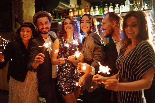 Group of happy people holding sparklers at the party. Young friends clinking glasses of champagne and enjoying new years eve with fireworks. Party, celebration, holidays concept.