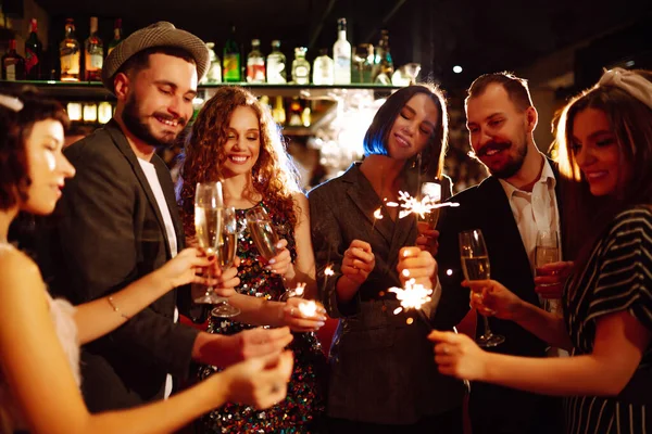 Group of happy people holding sparklers at the party. Young friends clinking glasses of champagne and enjoying new years eve with fireworks. Party, celebration, holidays concept.