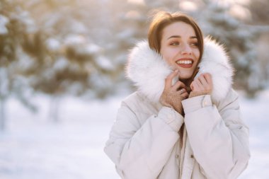 Kar parkında poz veren kışlık elbiseli güzel bir kadın. Ormanda güneşli bir kış gününde yürüyen genç bir bayan. Kış modası, tatiller, dinlenme, seyahat konsepti.