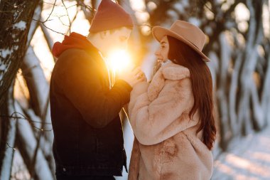 Genç çift kış tatilinde, gün batımında karlı bir ormanda. Kış günü kız arkadaşını kucaklayan yakışıklı bir genç. İnsanlar, mevsim, seyahat, aşk ve eğlence konsepti.