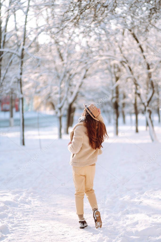 Stylish woman enjoying winter moments in a snowy park. Young woman in winter clothes posing with joy outside, frosty day. Youth, fashion, lifestyle and leisure concept.